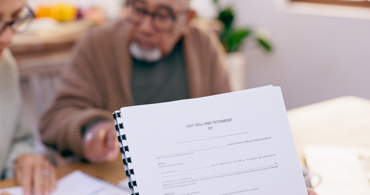 Document titled "Last Will and Testament" held by an individual during an estate planning meeting, with an older couple discussing estate planning details in a home setting.